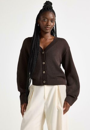 Pitkä lettipäinen nainen pukeutunut tummanruskeaan, napitettuun neuletakkiin ja korkeavyötäröisiin kermaisiin pliseerattuihin housuihin, seisoo yksivärisen taustan edessä.