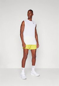 Vīrietis stāv, tērpies baltā bezroku Nike krekliņā, dzeltenās šortās, baltās Nike zeķēs un baltās sporta kurpēs, uz vienkrāsaina gaiša fona.