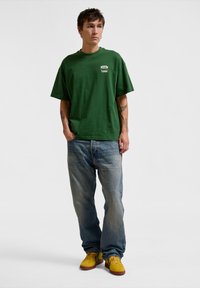 Green cotton T-shirt with a crew neck and short sleeves, paired with light blue denim jeans and yellow sneakers. Simple, relaxed fit.