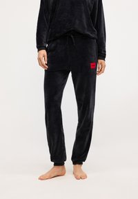 Čierny velúrový loungewear set s voľným strihom a tesnými nohavicami. Obsahuje červenú nášivku s logom na nohaviciach a elastické manžety. Mäkká textúra.