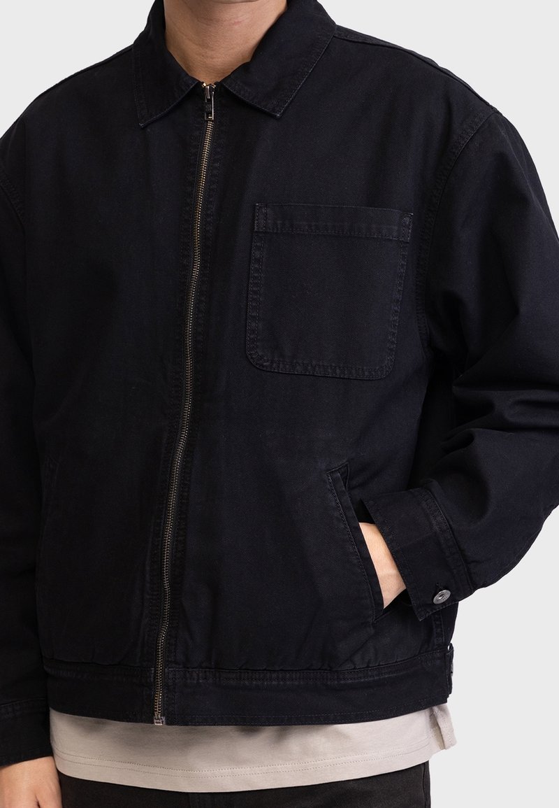 Homme portant une veste noire zippée avec une poche poitrine et des poches latérales, main dans la poche droite, par-dessus une chemise beige.