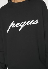 Czarna bluza wykonana z mieszanki bawełny, z białym, kursywnym logo "pequs" na piersi. Widoczne szczegóły ściągaczy.
