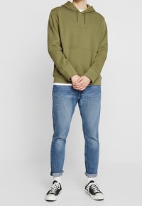 Persona che indossa una felpa verde oliva, jeans azzurri arrotolati, calzini bianchi e sneaker bianche e nere, in piedi con le mani giunte.