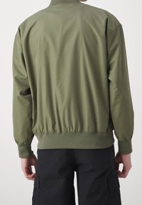 Giacca bomber verde oliva con vita e polsini elasticizzati, caratterizzata da una texture del tessuto liscia e un colletto alto.