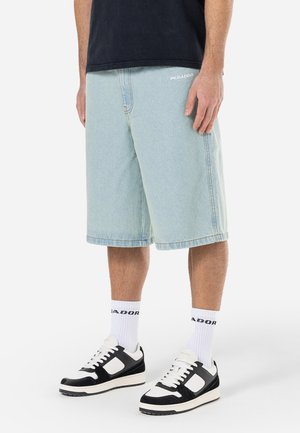 Mand iført lyseblå denimshorts, hvide sokker med sort logo, sorte og hvide sneakers og en sort skjorte, stående på en hvid baggrund.