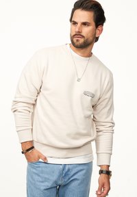 Beiger Sweatshirt mit rundem Halsausschnitt und langen Ärmeln. Mit einem kleinen gestickten Logo auf der Brust. Kombiniert mit hellblauen Jeans.