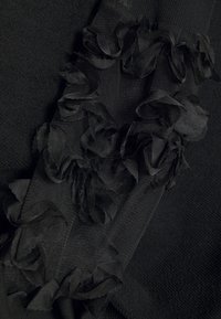 Tejido negro con punto texturizado y suaves acentos en volantes hechos de material transparente, creando un efecto visual y táctil en capas.