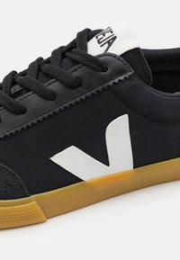 Melnas auduma sporta kurpes ar teksturētu melnu virsmeti, baltu V logotipu sānos un gumijas zoli. Iekļautas melnas šņores un balta valde uz mēles.