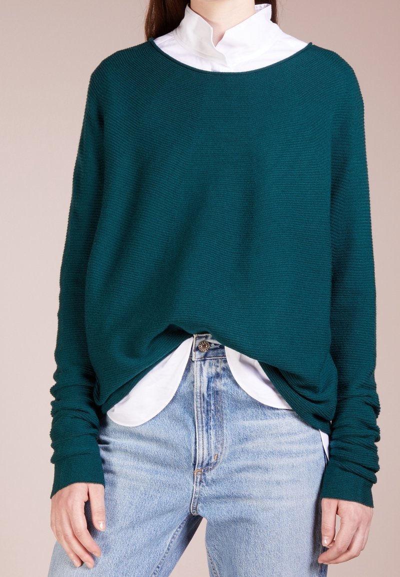 Personne portant un pull en tricot bleu sarcelle sur une chemise blanche non rentrée et un jean bleu clair, mains détendues sur les côtés, fond neutre.