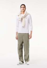 Hvid langærmet skjorte, olivengrøn cargo-bukser, beige kabelstrikket sweater hængende over skuldrene, og hvide sneakers med grønne detaljer.