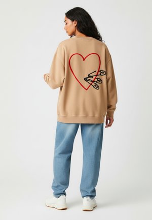 Felpa beige con un design a forma di cuore rosso e spille da balia nere sul retro, abbinata a jeans blu chiaro e sneaker bianche.