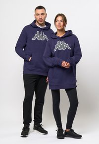 Dos personas llevan sudaderas moradas con un gran logotipo blanco. El hombre tiene un bolsillo frontal, mientras que la sudadera de la mujer es más larga. Ambos llevan pantalones negros.