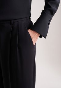 Zwarte op maat gemaakte broek met een gladde textuur, met plooien aan de voorkant en een zijzak, gecombineerd met een donker, lang mouwen shirt.