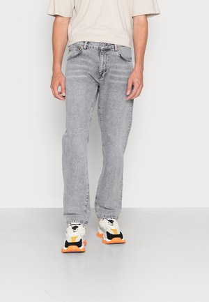 Persoon draagt lichtgrijze verweerde jeans en witte sneakers met oranje zolen, staand tegen een effen witte achtergrond.