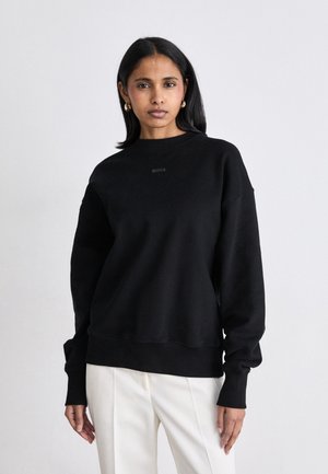 Felpa nera con scollo a girocollo e spalle basse, con un discreto logo "BOSS". Tessuto liscio, vestibilità ampia, abbinata a pantaloni bianchi.