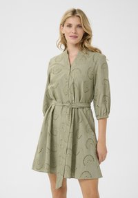 Grön knälång klänning med knapplist fram, 3/4-långa ärmar och smal midja. Har broderade spiraler och ögla utskärningar för textur.