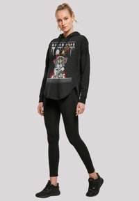 Fekete kapucnis pulóver grafikus mintázattal, amely karikatúra karaktereket ábrázol, bordázott mandzsettákkal és ívelt aljjal. Fekete leggings-szel és sportcipővel párosítva.