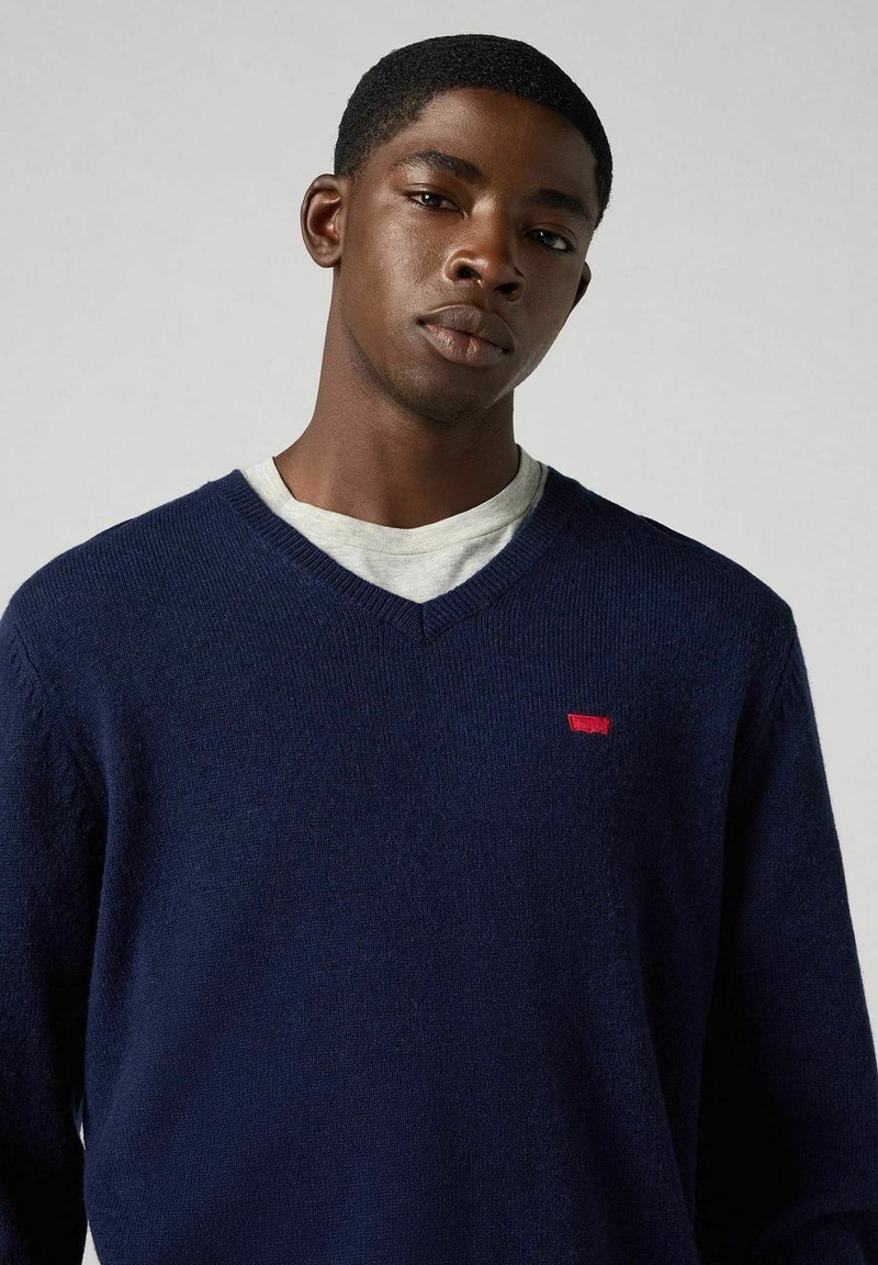 Marineblauer Wollpullover mit V-Ausschnitt-Design. Besitzt ein kleines rotes Logo auf der Brust und ein strukturiertes Strickmuster. Darunter befindet sich ein hellgraues T-Shirt.