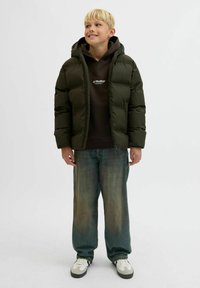 Dreng smilende, iført en sort dunjakke, sort hættetrøje med logo, løse blå jeans og hvide sneakers, stående mod en ensfarvet baggrund.