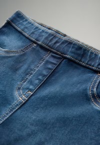 Mėlyni džinsai iš denimo su kontrastingais siuvinėjimais ant juosmens, priekinėmis kišenėmis ir lygia tekstūra. Dėmesys detalizuotiems siūlams ir apdorojimui.