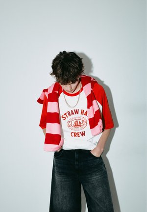 Jeune personne aux cheveux foncés regardant vers le bas, portant un t-shirt rouge et blanc "Straw Hat Crew", un pull rayé rouge et rose, et un jean foncé.