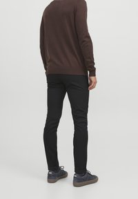 Homme portant un pull marron à manches longues, un pantalon noir slim et des baskets foncées, debout, dos à un fond uni.