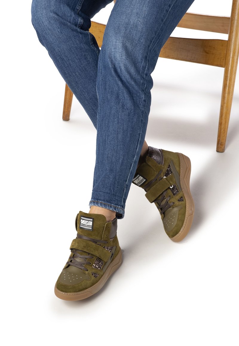 Personne assise sur une chaise en bois portant un jean bleu et des baskets montantes vertes avec une semelle gomme et des sangles à Velcro.