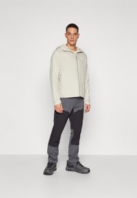 Letvægts, beige zip-jacket med hætte, parret med tætsiddende, mørkegrå bukser med lysere grå detaljer og sorte sko.