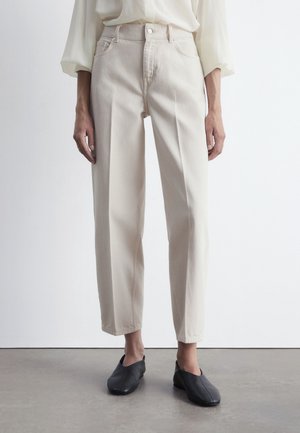 Beżowe spodnie z wysokim stanem i prostymi nogawkami, z zapięciem na guzik z przodu oraz kieszeniami bocznymi. W połączeniu z czarnymi butami wsuwanymi.