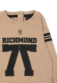 Felpa beige con scritta nera "RICHMOND" e grande "77" sul davanti. Righe nere sulle maniche, collo a coste con chiusure a scatto. Morbido materiale a maglia.