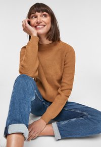 Bruine geribbelde trui met een hoge hals en textuursleeves, gedragen met lichtblauwe denim met opgerolde broekspijpen, zittend tegen een effen achtergrond.