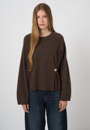 Kobieta o długich blond włosach, ubrana w luźny brązowy sweter ze złotym guzikiem i ciemnoniebieskie jeansy, stojąca na jednolitym tle.