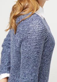 Femme aux longs cheveux blonds portant un pull en maille texturée bleu par-dessus une chemise blanche à col, vue de profil sur un fond uni.