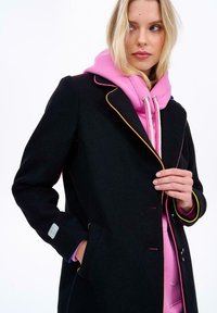 Blondínka v čiernom kabáte so farebnými lemami cez ružovú mikinu, pozerajúca sa na stranu a jednou rukou si upravujúca kabát.