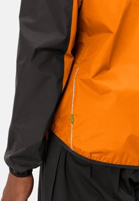 Giacca a vento con pannelli arancioni e neri, materiale leggero, polsini elastici e un accento blu a contrasto lungo la cucitura.
