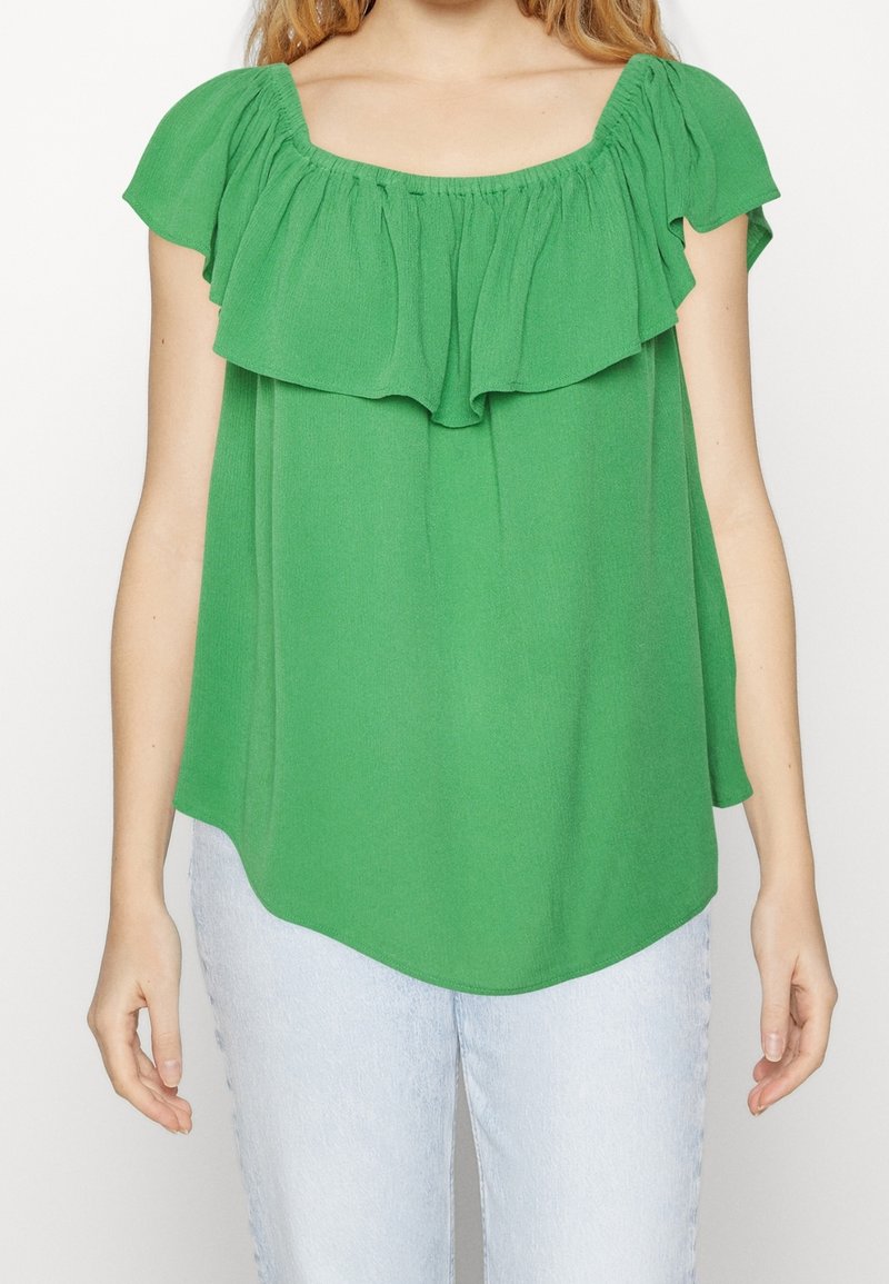 Femme portant un chemisier vert vif sans épaules avec un col volanté et un jean bleu clair, debout devant un fond blanc uni.