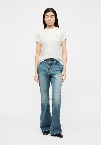 Γυναίκα που στέκεται φορώντας λευκό κοντομάνικο μπλουζάκι με λογότυπο και ψηλόμεσα καμπάνες μπλε τζιν με μαύρα παπούτσια σε λευκό φόντο.