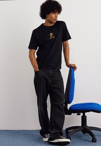 Schwarzes T-Shirt mit kurzen Ärmeln und kleinem goldenen Logo. Getragen mit dunklen Jeans und schwarz-weißen Sneakers. Hintergrund mit einem blauen Drehstuhl.