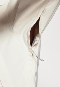Beige Jacke mit seitlichem Reißverschluss und verstellbarem Kordelzug; glatter Stoff mit subtiler Textur und verstärkten Nähdetails.