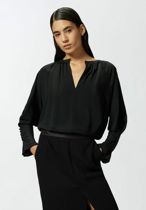 Blusa negra de corte holgado con escote en V, cuello fruncido y mangas largas; tela elegante con ligero brillo y detalles de botones en las mangas.