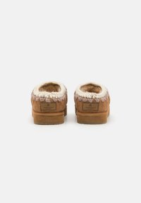 Pantofole in suede marrone con rivestimento in shearling bianco, dettagli di cucitura circolare attorno all'apertura e suola piatta in gomma. Logo del marchio visibile sul retro.