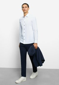 Weißes Langarmhemd mit dunklen Knöpfen, kombiniert mit marineblauen Hosen. Ein marineblauer Blazer wird am rechten Arm getragen, und weiße Sneaker vervollständigen den Look.