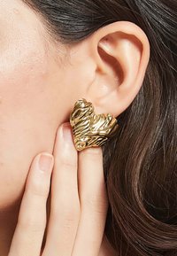 Guldörhänge i hjärtform med en strukturerad, vågig yta, uppvisad mot en närbild av en persons öra och hand.