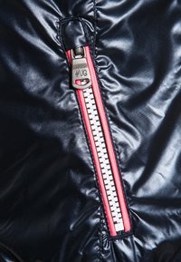 Chaqueta acolchada de un azul marino oscuro con una textura brillante, presenta un cierre rojo prominente y herrajes plateados con una etiqueta que dice "#UG".