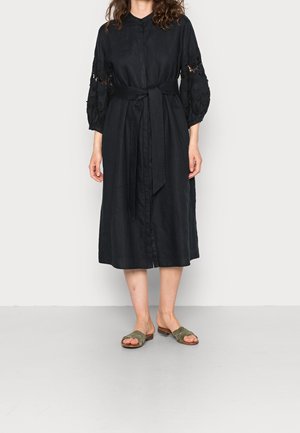 Femme portant une robe midi noire avec une ceinture et des détails en dentelle sur les manches, associée à des sandales plates ouvertes vert olive.