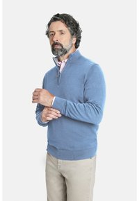 Suéter de cuello con cremallera en un suave tejido de color azul claro. El diseño presenta puños y cintura de canalé, usado sobre una camisa rosa con cuello.