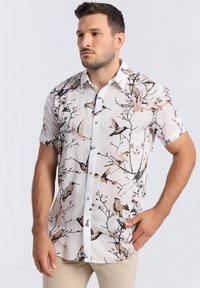 Chemise à manches courtes de couleur blanche avec un motif floral et d'oiseaux, dotée de boutons et d'un col classique. Le matériau semble léger et texturé.