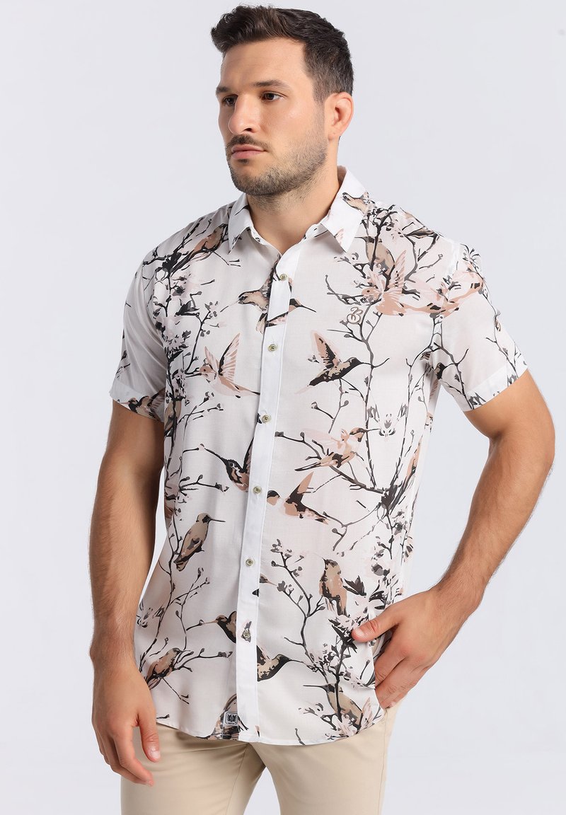 Chemise à manches courtes de couleur blanche avec un motif floral et d'oiseaux, dotée de boutons et d'un col classique. Le matériau semble léger et texturé.