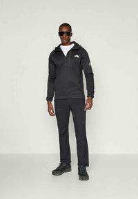 Mand iført sort North Face hættejakke, sorte bukser, sorte vandresko og mørke solbriller, stående mod en ensfarvet lys baggrund.