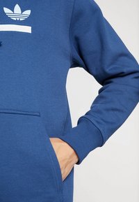 Sweatshirt bleu avec un logo blanc, une poche avant, des poignets côtelés et un tissu texturé. Gros plan sur la main dans la poche.